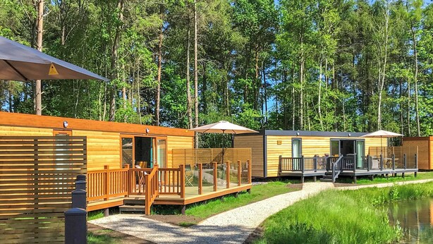A row of wooden cabins with outdoor seating and a gravel path leading to a body of water. - Cedar Boutique Lodge (Aughton)