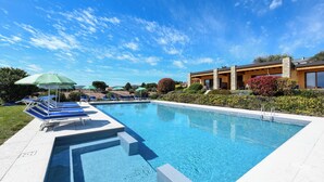 Une piscine rectangulaire à l’eau d’un bleu limpide, entourée d’une terrasse blanche. On y trouve des chaises longues bleues et un parasol vert sur le côté gauche.