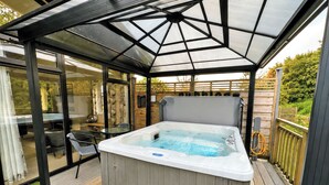 Un bain à remous installé sous une terrasse fermée par des baies vitrées, avec une plateforme en bois et un petit banc.