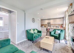 A modern living room with green sofas, a wooden coffee table, and a kitchen in the background.