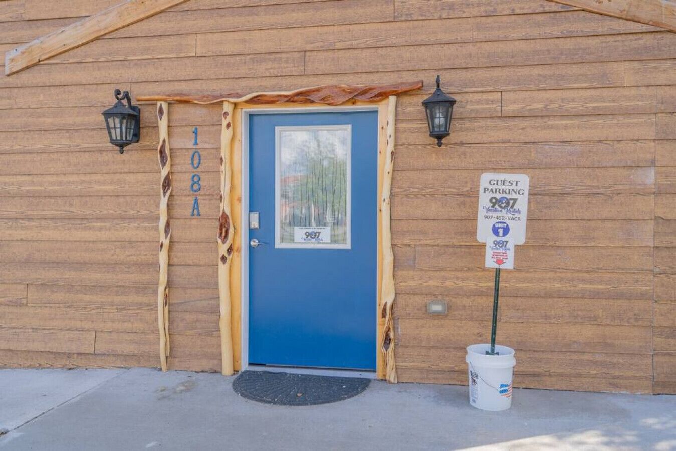 Un bâtiment en bois avec une porte bleue, portant le numéro 108A, et deux lanternes noires de chaque côté.
