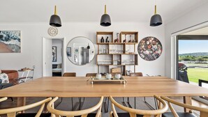 Een eethoek met een houten tafel, stoelen, hanglampen, een spiegel, planken met glazen en uitzicht door een raam.