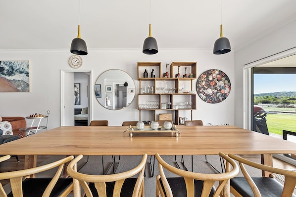 A dining area with a wooden table, chairs, pendant lights, a mirror, shelves with glassware, and a view through a window.