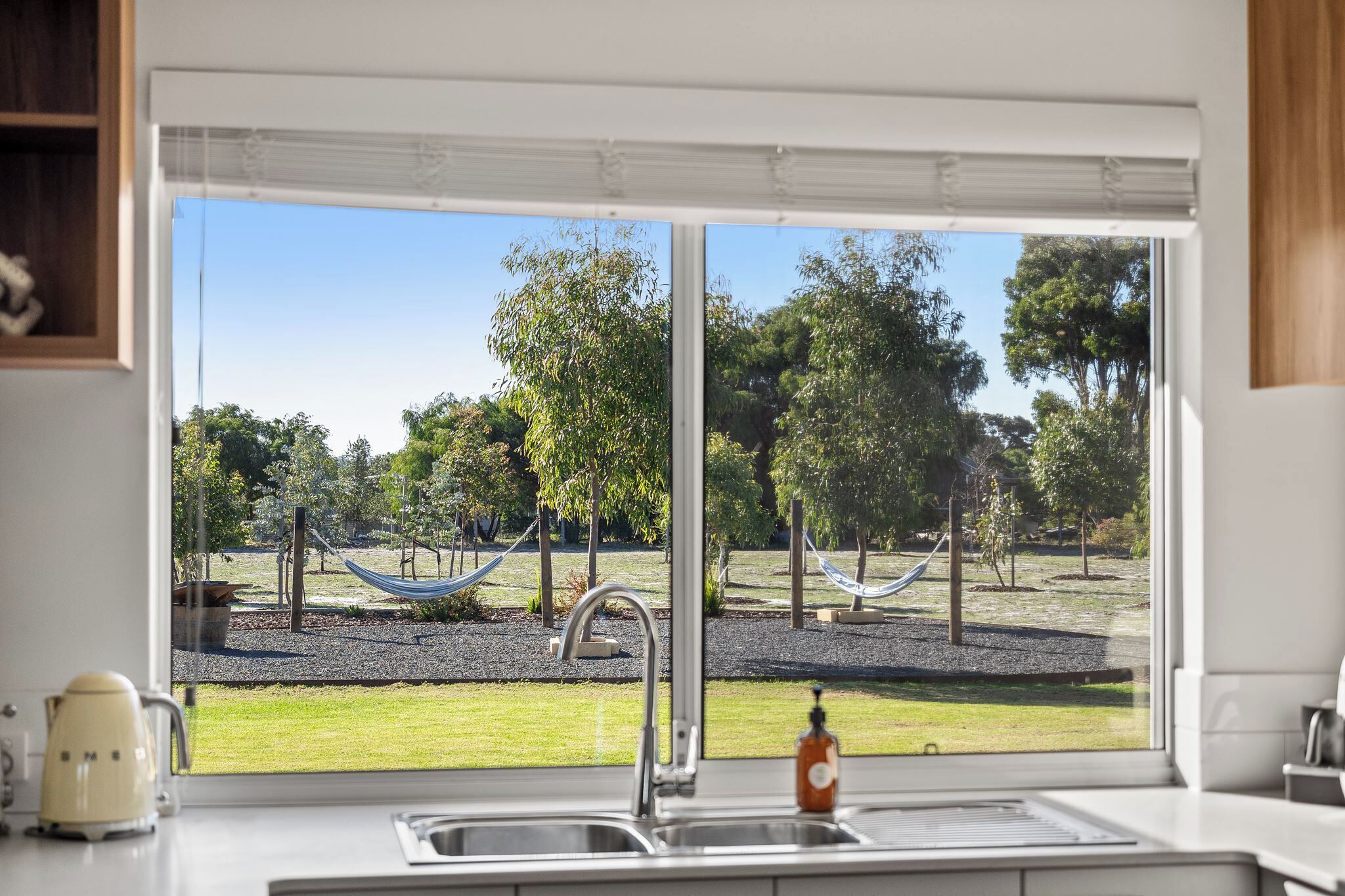 Una cocina con un ventanal grande, un lavamanos y vistas a un parque con árboles y una hamaca.