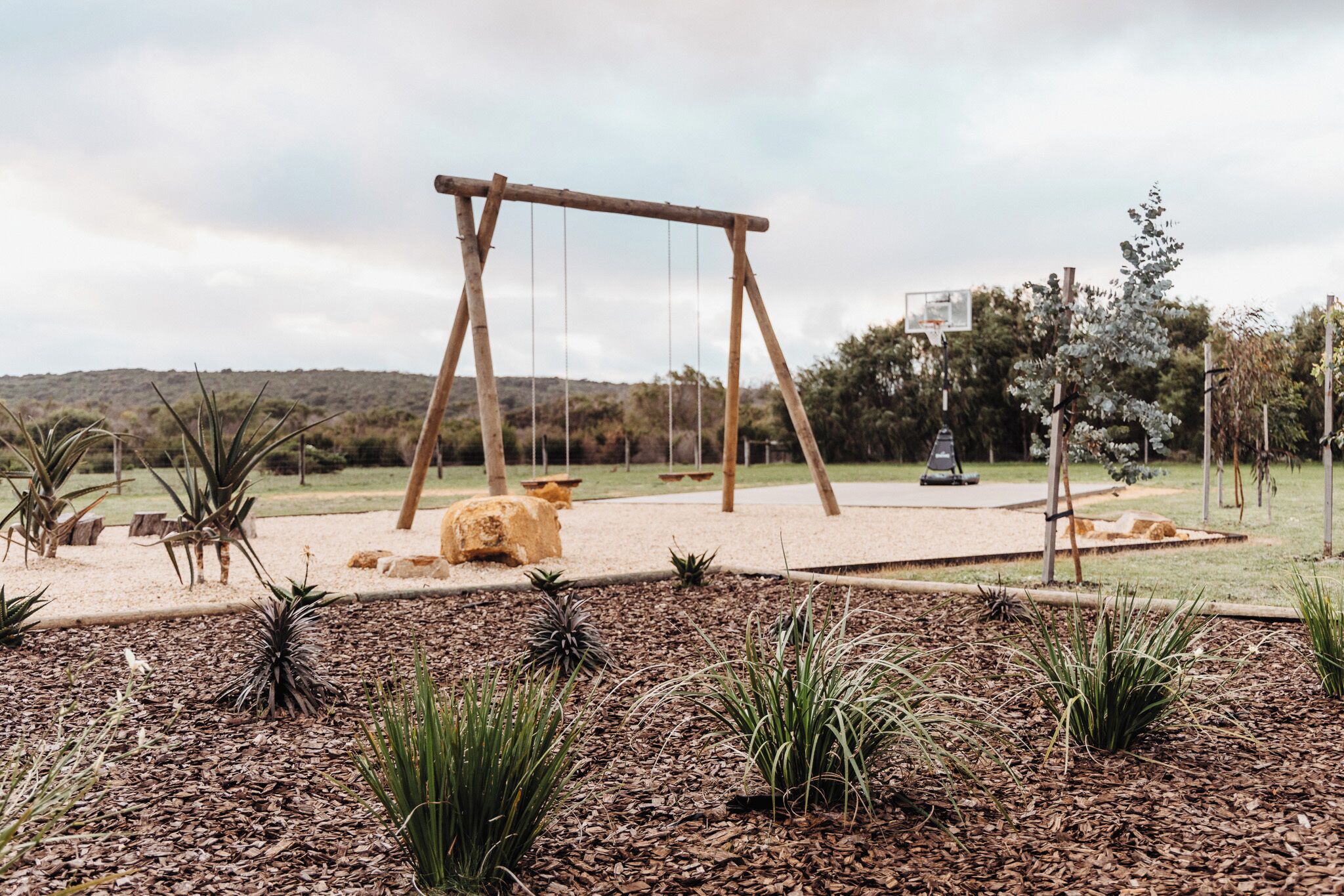 Un parc équipé d’une balançoire en bois, d’un terrain de basket et de diverses plantes.