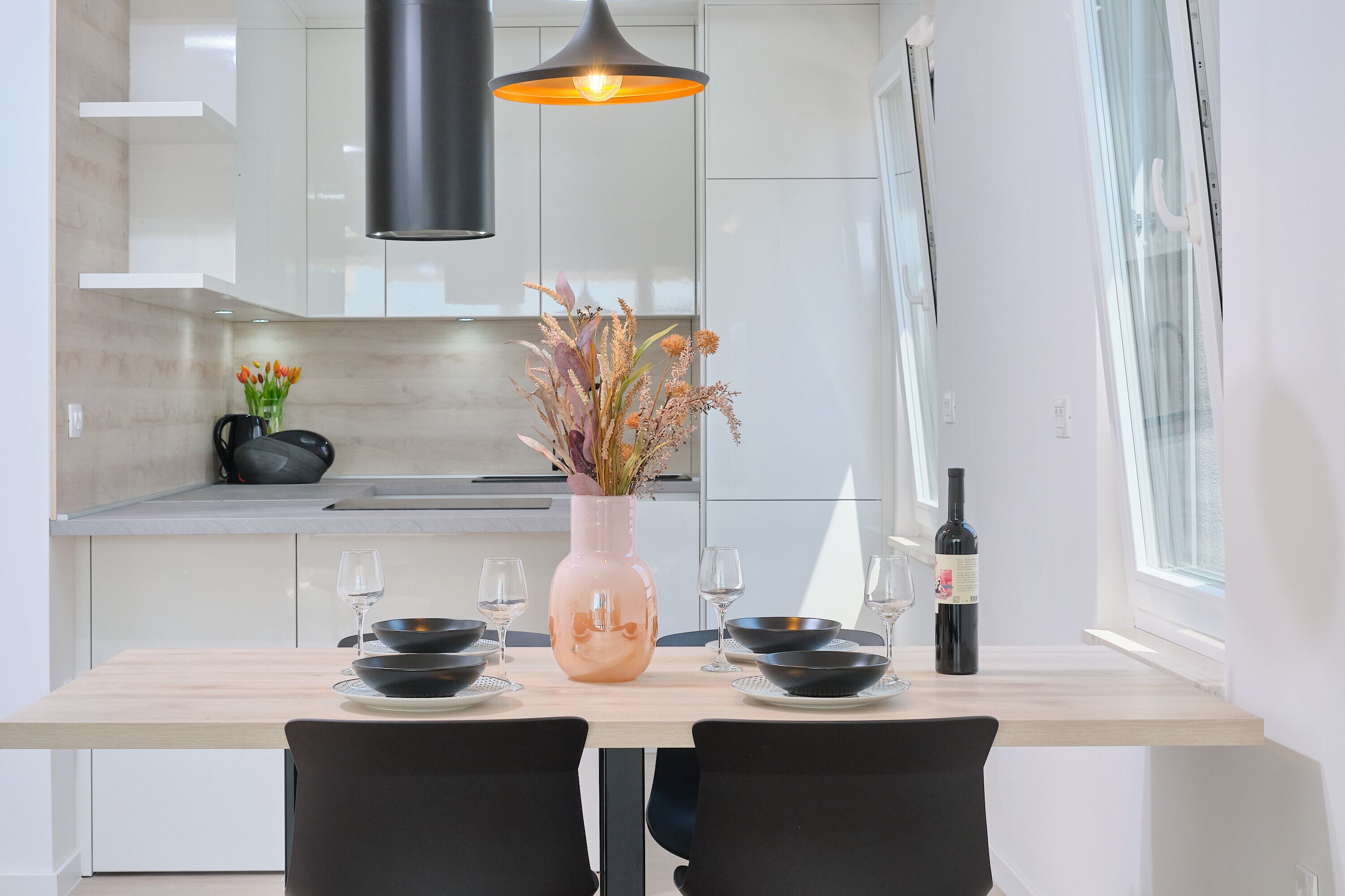 A modern kitchen with white cabinets, a wooden dining table, black chairs, and a vase with flowers.