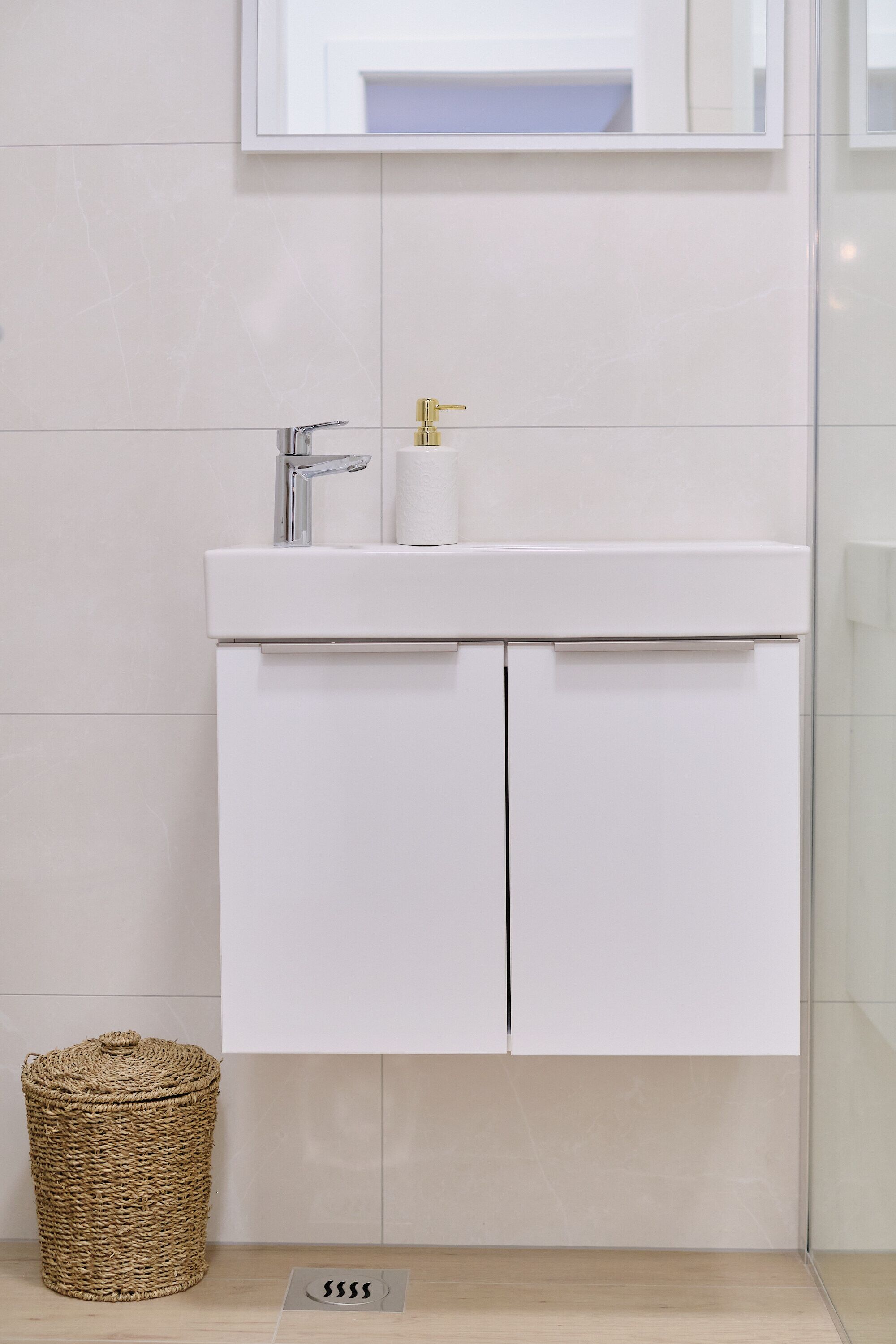 A modern bathroom with a white sink, chrome faucet, and a single roll of toilet paper. A woven basket is placed on the floor beside the sink.