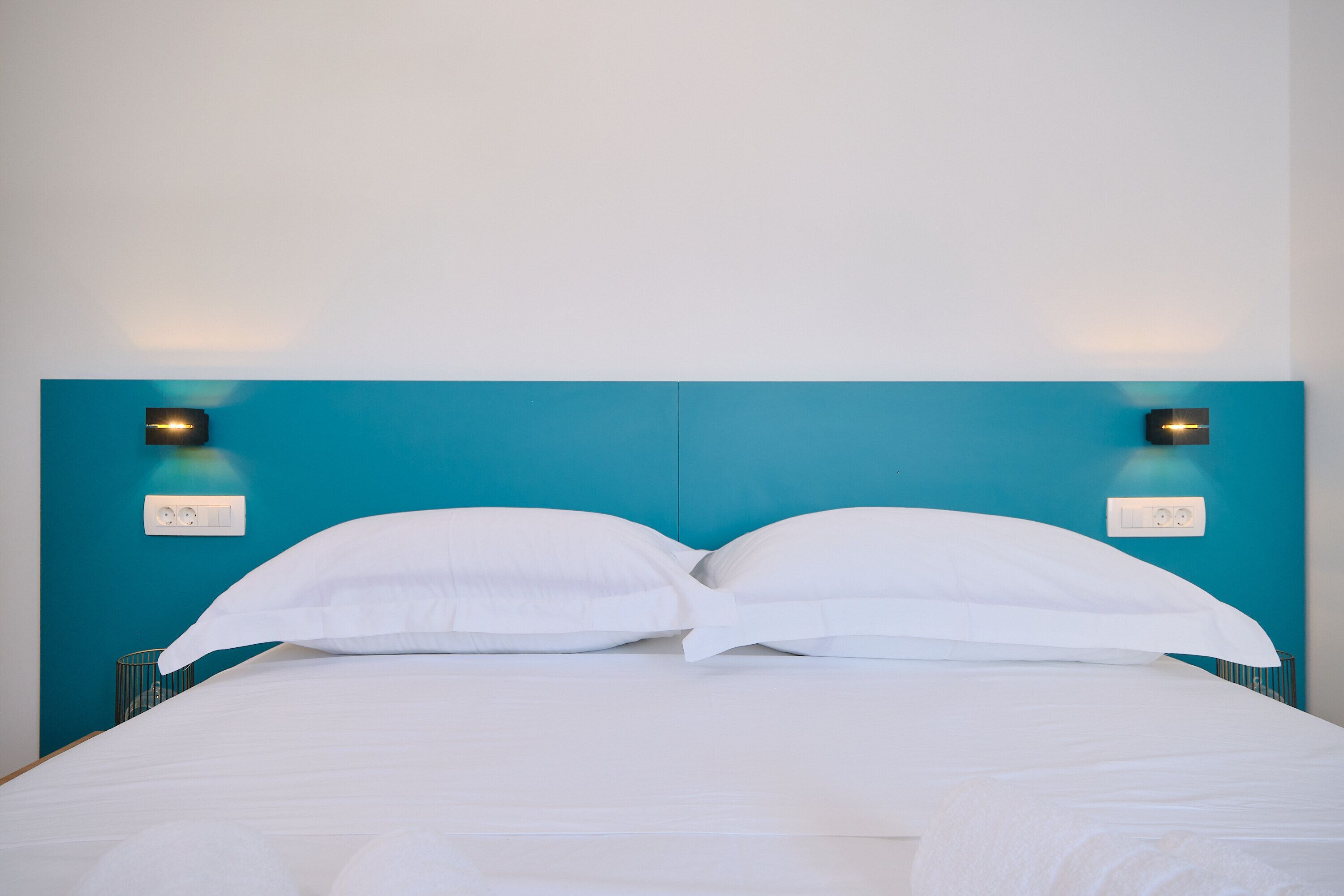 Un lit avec une literie blanche, une tête de lit de couleur turquoise et deux appliques murales.