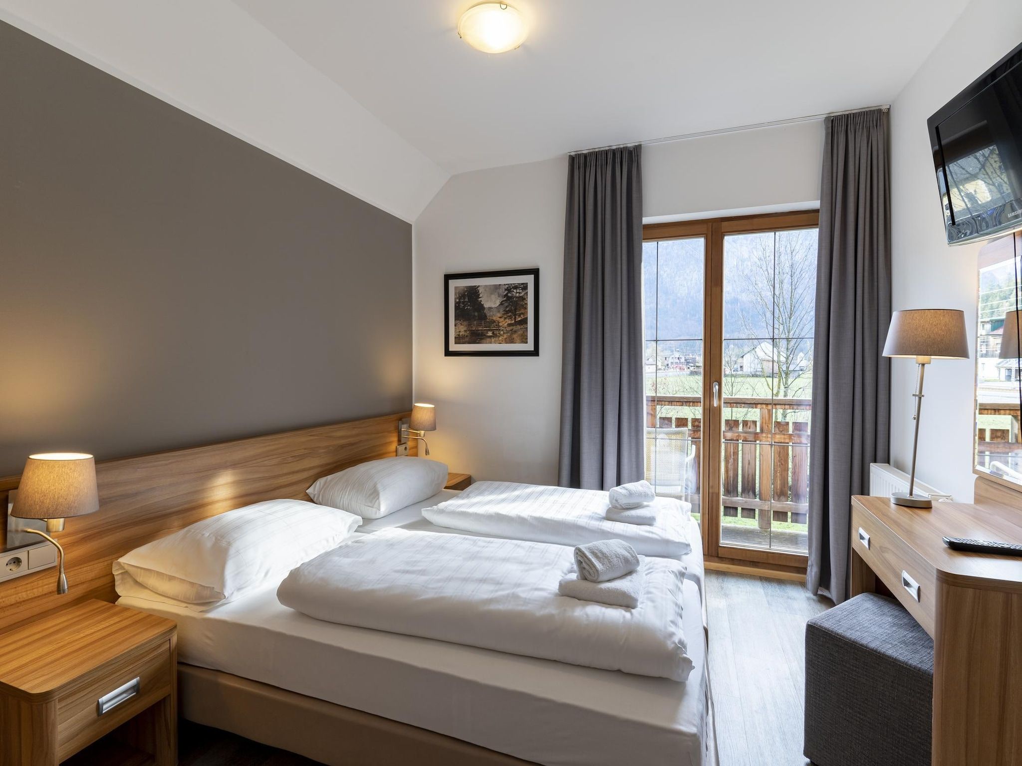 Habitación de hotel con cama, lámparas de noche, escritorio con silla, televisor y ventana con cortinas.