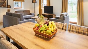 Una camera d'albergo con un tavolo da pranzo in legno, un cesto di frutta, un divano, una televisione e una finestra con tende.