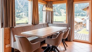 Een eetgedeelte met een houten tafel en een gewatteerde bank, grote ramen met gordijnen en uitzicht op een bergachtig landschap.