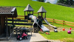 Un terrain de jeux avec un toboggan, un petit bâtiment et une zone pavée où se trouve une voiture jouet.