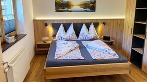 Une chambre à coucher moderne avec un lit en bois, une table de chevet, une commode, une fenêtre avec des stores, un radiateur et un tableau représentant un paysage.