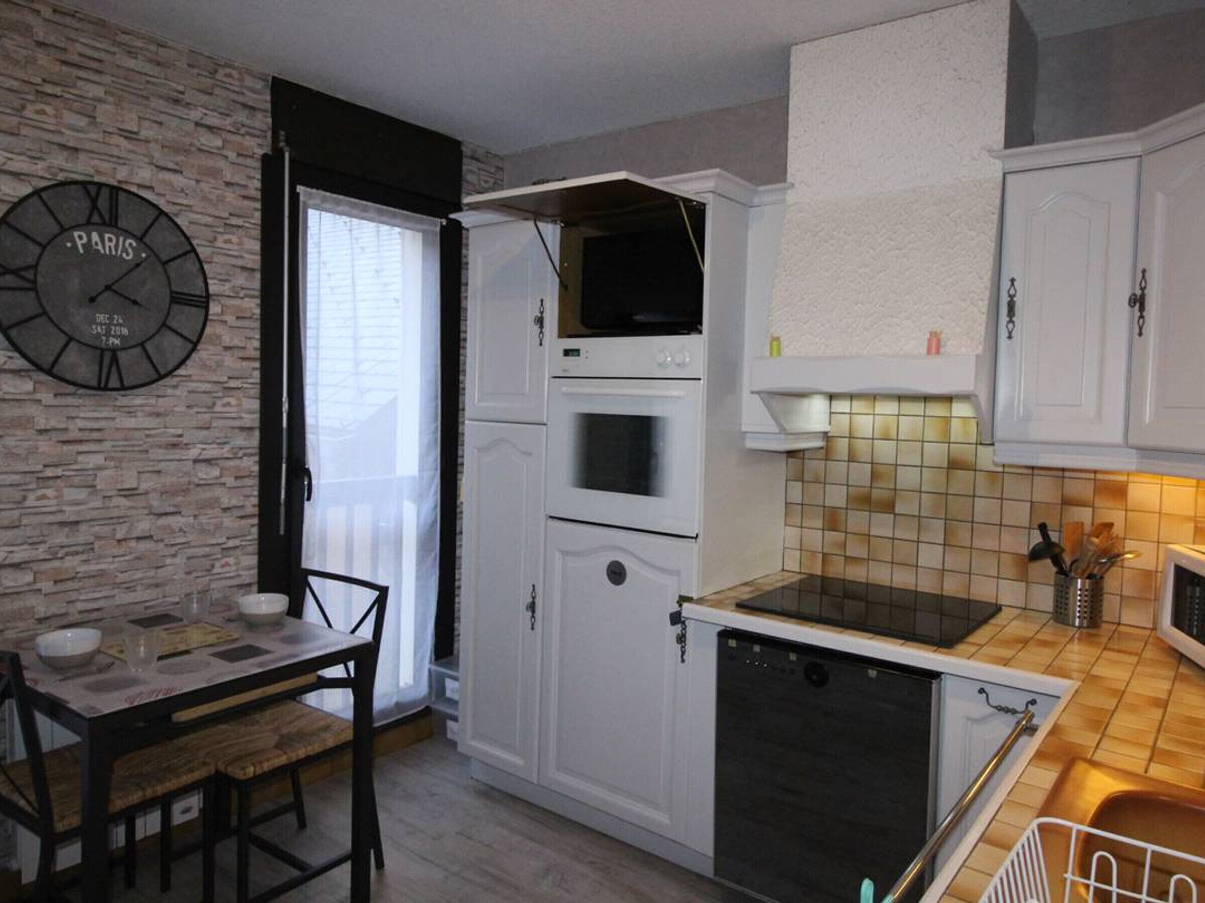Een kleine keuken met witte kasten, een inbouwoven, een magnetron, en een eethoek met een tafel en stoelen.