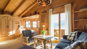 En mysig stuga med träpanel på insidan, med ett matområde, en soffa och utsikt över bergen genom de stora fönstren.