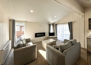 A modern living room with a flat-screen TV, two sofas, a coffee table, and a large window with curtains. - Superior Lodge (sleeps 8) (Bacton-on-Sea)