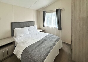 A bedroom with a bed, bedside table, and a window with curtains. - Superior Lodge (sleeps 8) (Bacton-on-Sea)
