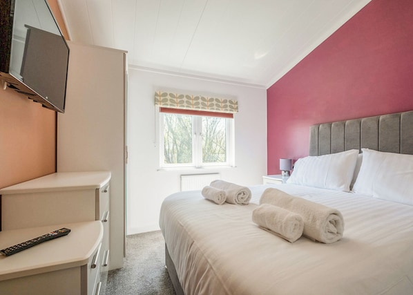 A bedroom with a bed, a TV mounted on the wall, a bedside table, and a window with a view of trees.