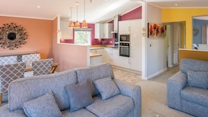 A modern living room with a grey sofa, a dining area with patterned cushions, and a kitchen with white cabinets and a red accent wall.