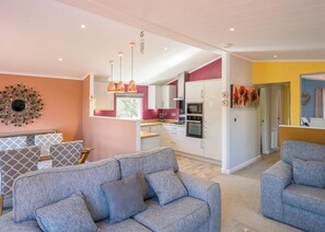 A modern living room with a grey sofa, a dining area with patterned cushions, and a kitchen with white cabinets and a red accent wall. - 3 bedroom accommodation in Carnforth (Carnforth)