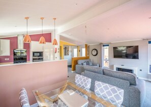 A modern living room with a sofa, a glass coffee table, pendant lights, a kitchen area, and a TV mounted on the wall. - 3 bedroom accommodation in Carnforth (Carnforth)