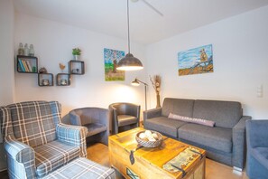 Una sala de estar con sofá, sillón y una mesa de centro con un cuenco de pan. Se observan estantes en la pared con objetos decorativos y dos cuadros.