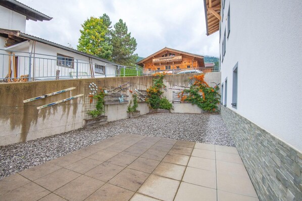 Un patio con piso de baldosas, un muro de concreto con una planta trepadora y un edificio de madera al fondo.