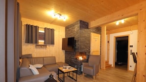 A cozy living room with a stone fireplace, wooden walls, a sofa, a coffee table, and a TV mounted on the wall. - Comforting chalet with whirlpool (Sankt Georgen am Kresichberg)