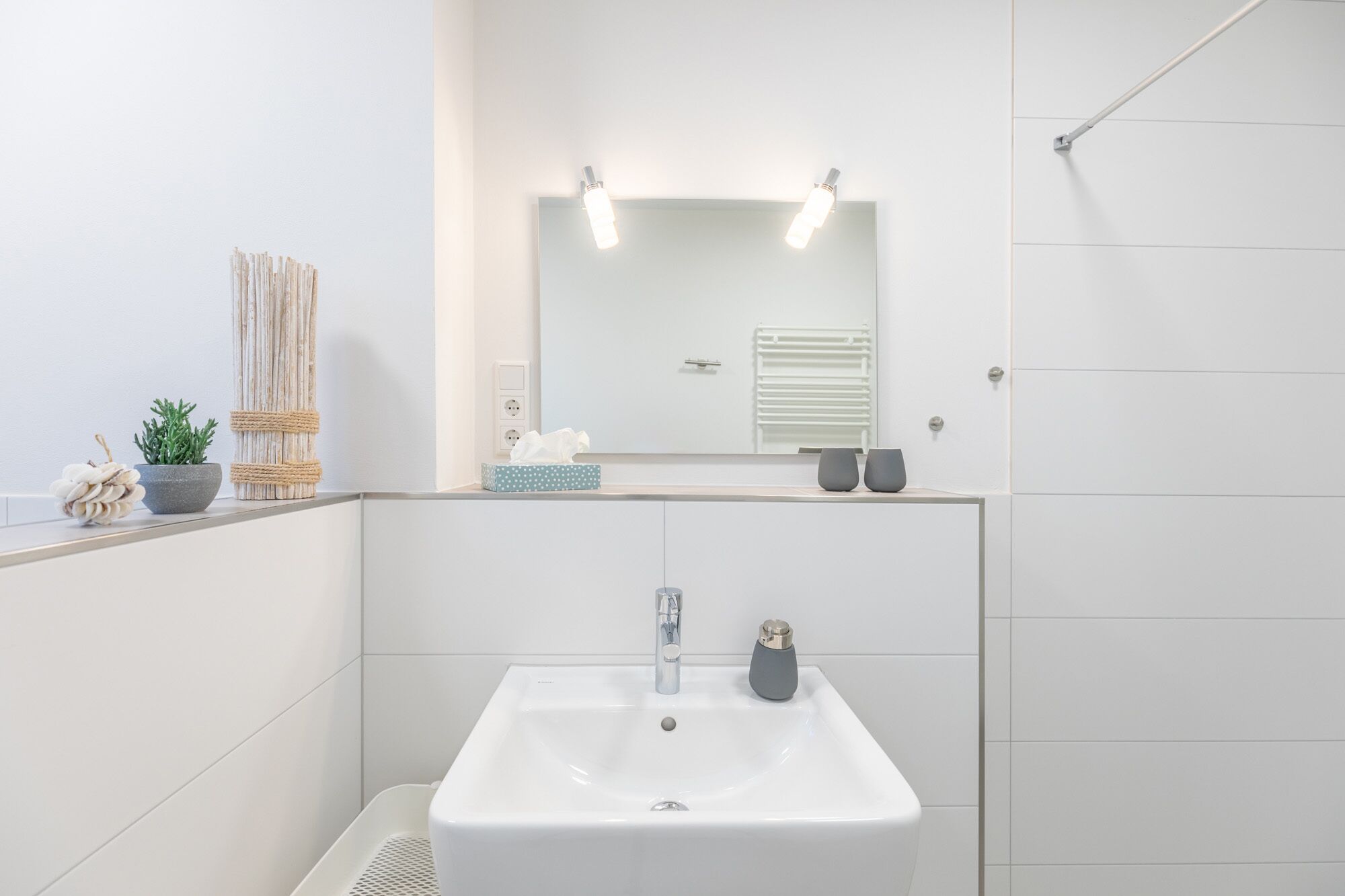Un baño moderno con un lavamanos blanco, un espejo con iluminación, un perchero para toallas y una pequeña planta.