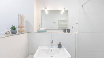 Un baño moderno con un lavamanos blanco, un espejo con iluminación, un perchero para toallas y una pequeña planta.