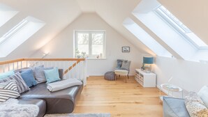 A cozy loft bedroom with a sloped ceiling, wooden flooring, a brown leather sofa, a small chair, a side table with a lamp, and a window with a view of trees.