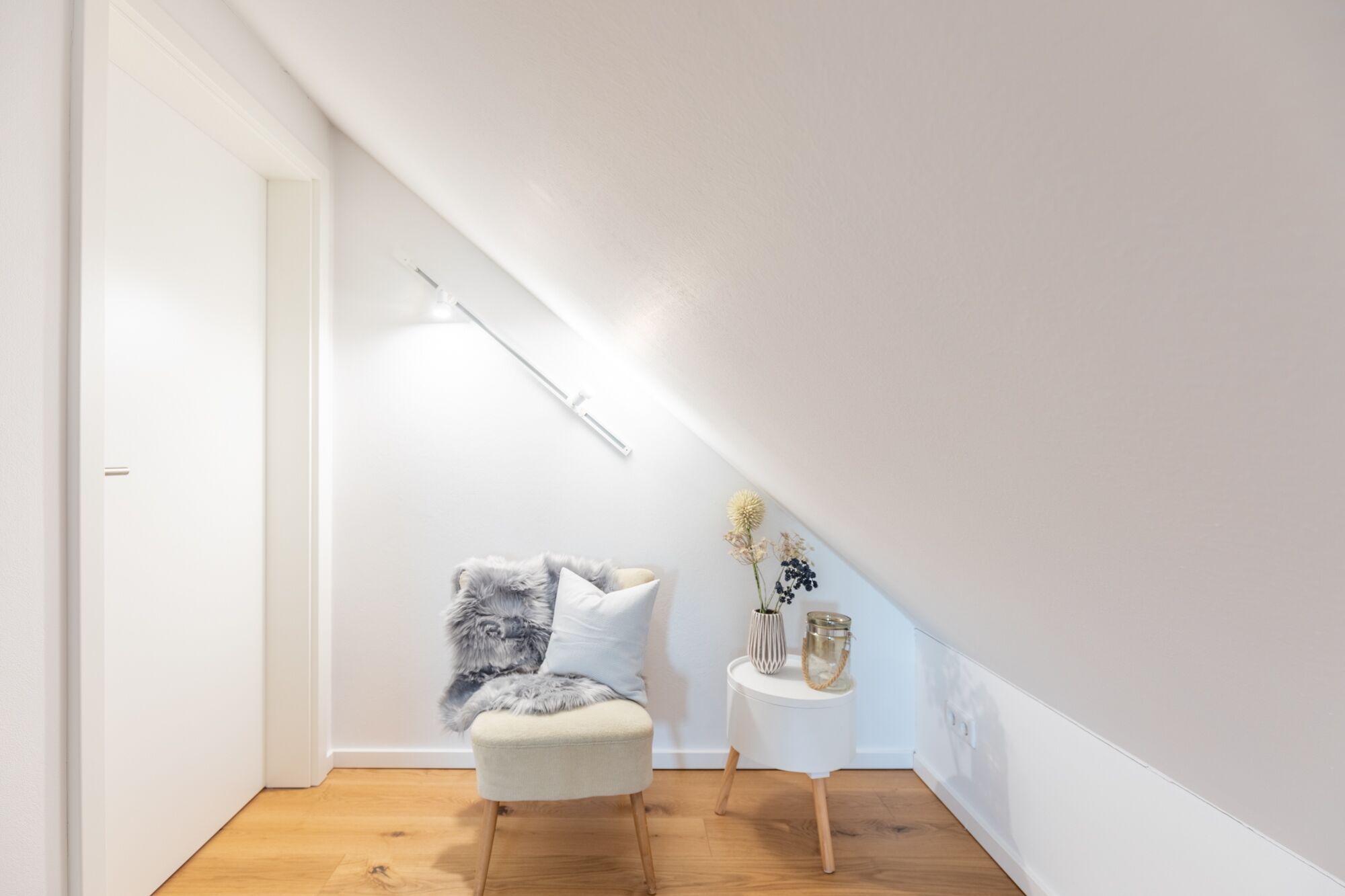 Une petite pièce bien éclairée, avec un plancher en bois, un fauteuil moelleux, une petite table d’appoint agrémentée d’objets décoratifs et un plafond en pente.