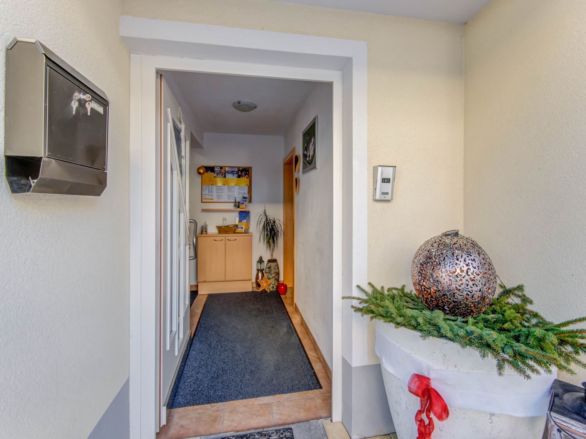 En hall med en dörr som leder till ett kök, en dekorativ vägglampa och en krukväxt med tallgrenar.