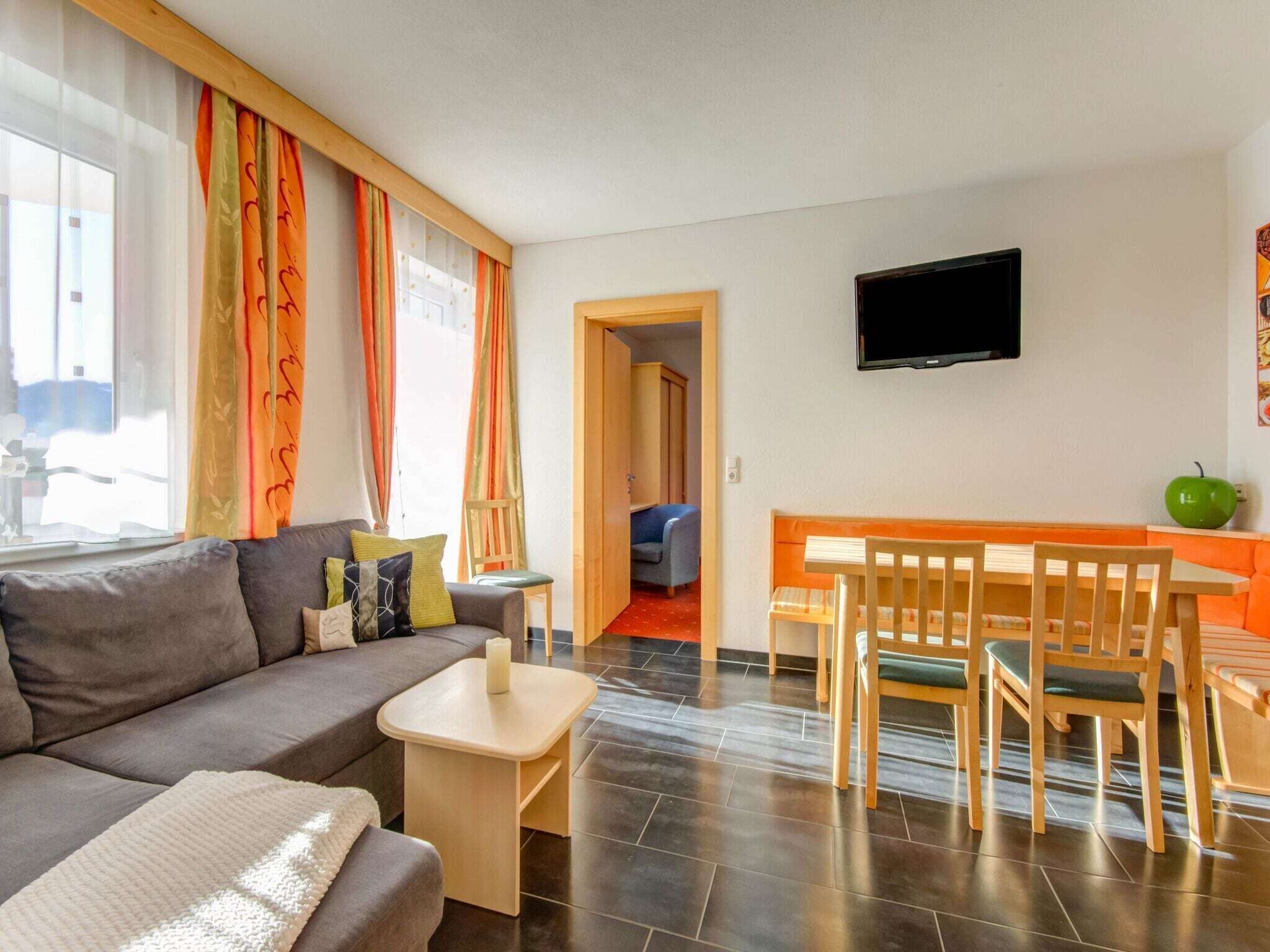 A modern living room with a sofa, a small table, and a dining area with chairs and a table. A television is mounted on the wall.