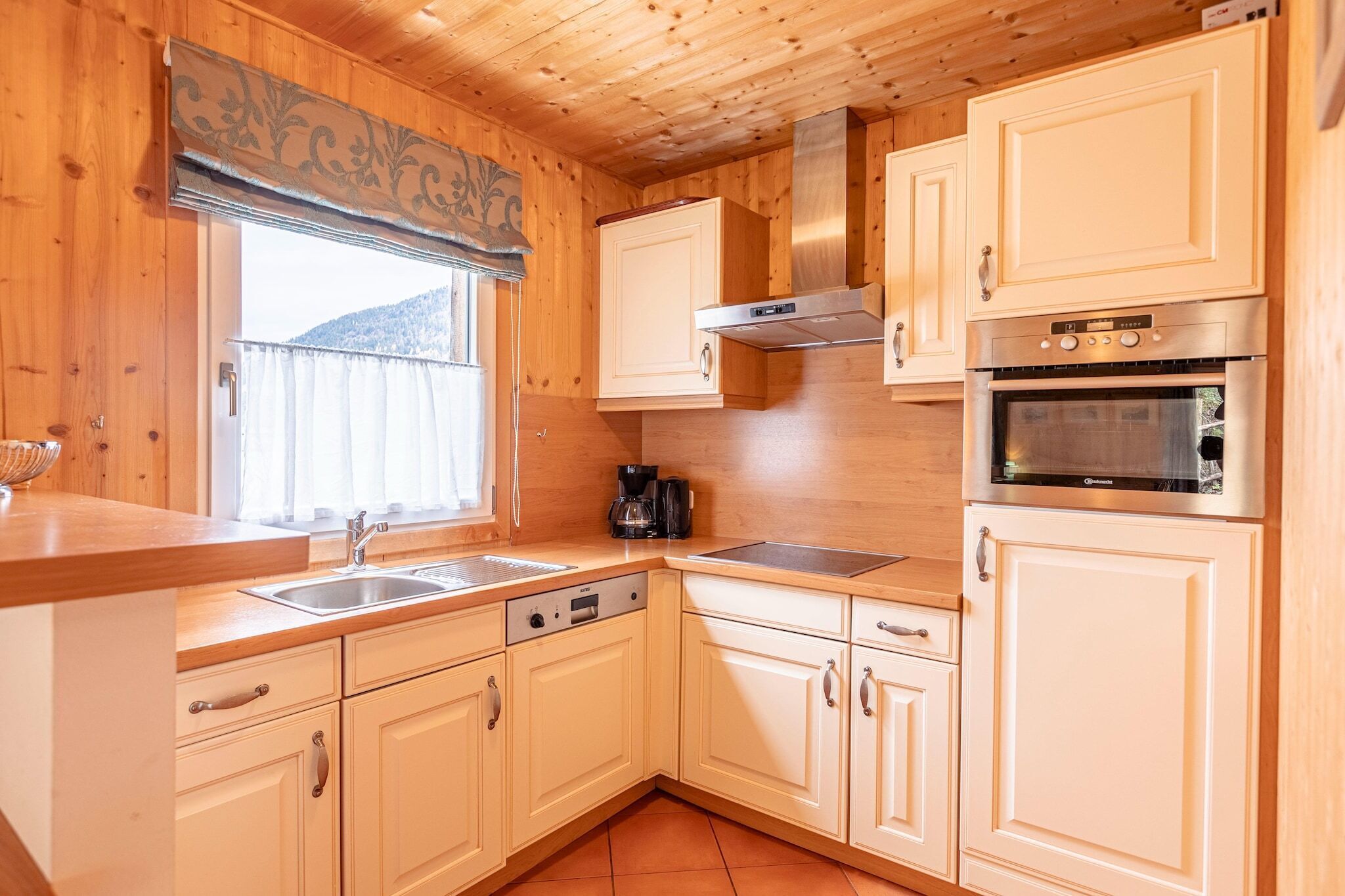 Eine Küche mit Holzschränken, einer Edelstahl-Spüle, einem Einbauofen und einem Fenster mit Vorhängen.