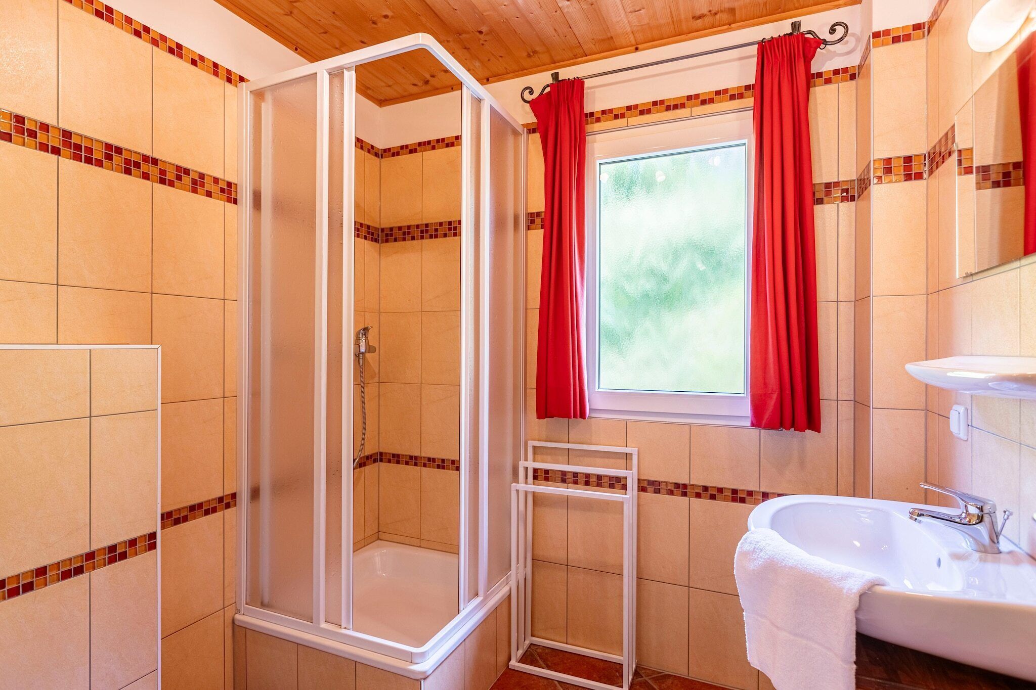 Ein Badezimmer mit Duschkabine, einem Fenster mit roten Vorhängen und einem Waschbecken mit Handtuch.