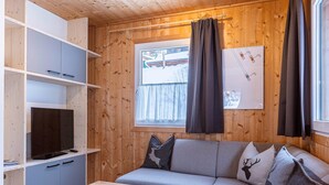 A cozy cabin interior with a wooden ceiling, a sofa with deer-patterned pillows, a TV on a shelf, and a window with curtains.