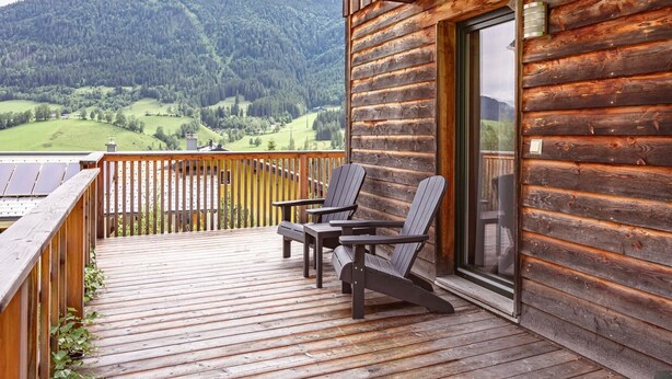 A wooden cabin with a deck, two Adirondack chairs, and a glass door leading to a balcony.
