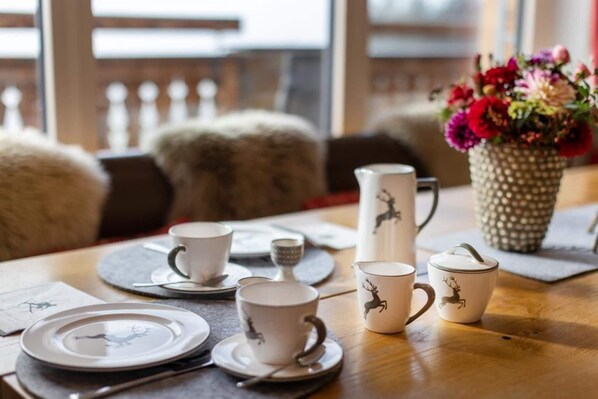 A dining table set with white cups and saucers, a teapot, and a pitcher, with a vase of flowers in the background.