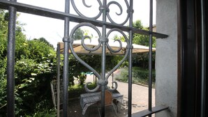 Blick durch ein Metallgitter auf eine Patio-Anlage mit Tisch und Stuhl, umgeben von viel Grün.