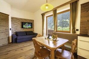 Un salon chaleureux avec un plancher en bois, un canapé foncé, une table à manger en bois avec des chaises, un téléviseur fixé au mur et une fenêtre avec des rideaux.