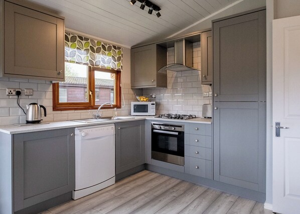 A modern kitchen with grey cabinets, a dishwasher, a microwave, a stove, and a window with a leaf-patterned curtain.