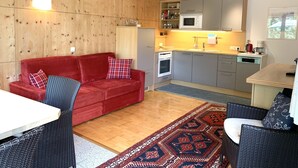 Un espai de vida compacte amb un sofà vermell, una taula de dinar, cadires de vimetla, una zona de cuina amb electrodomèstics moderns i una alfàbrica amb disseny.