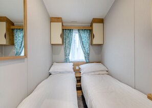 Um quarto pequeno e estreito, com duas camas, um espelho e uma janela com cortinas.