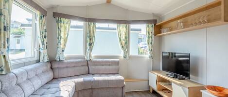 A modern living room with a curved sofa, a flat-screen TV, wooden shelves, and large windows with curtains.