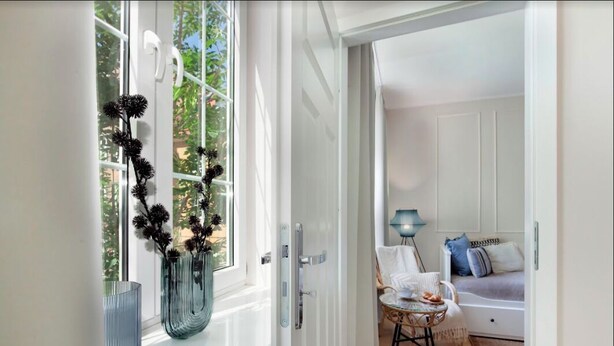 A room with a bed, a chair, a small table, and a window with a decorative plant.