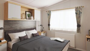 Een keurig opgemaakt slaapkamer met een bed, nachtkastjes, een plank met decoratieve objecten, een raam met gordijnen en een radiator.