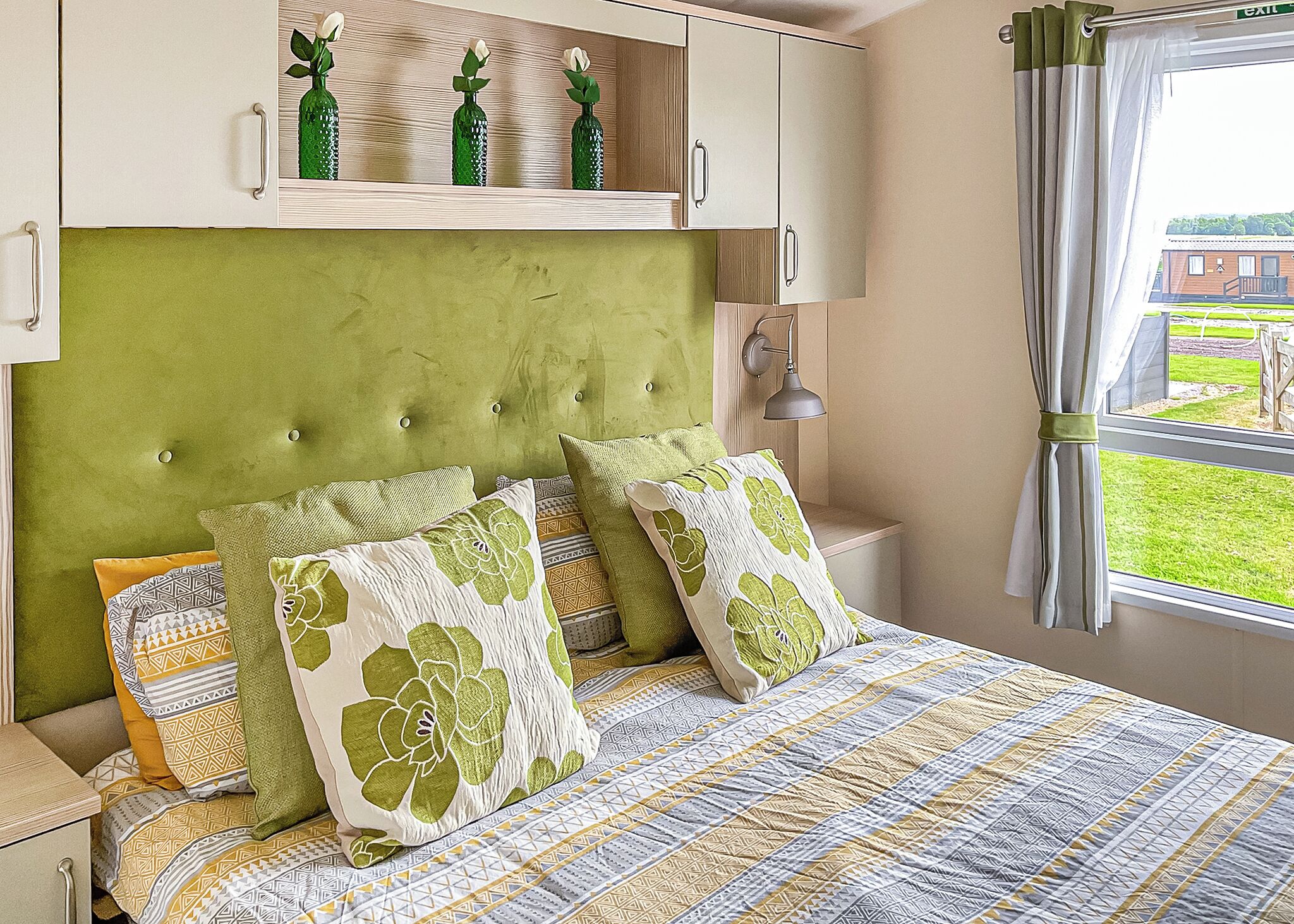A bedroom with a green upholstered headboard, a patterned bedspread, and a window with a view of a building and greenery.