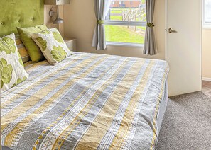 A bedroom with a bed, a green headboard, a window with curtains, and a view of a garden.