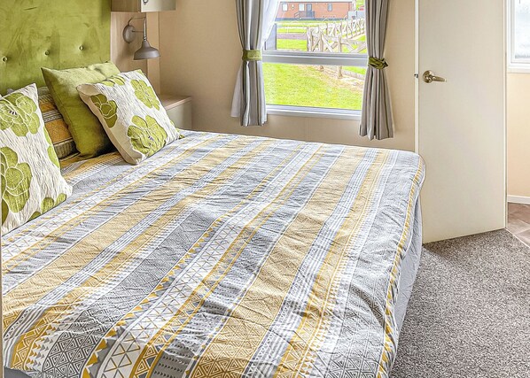 A bedroom with a bed, a green headboard, a window with curtains, and a view of a garden.